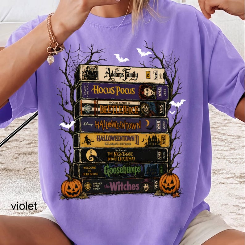 Op de afbeelding: Een violet t-shirt met een grafische print van een stapel van acht vintage VHS-banden met de titels The Addams Family, Hocus Pocus, Beetlejuice, Halloweentown, Halloweentown II, The Nightmare Before Christmas, Goosebumps en The Witches.