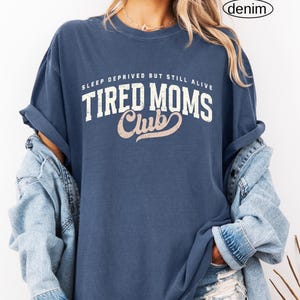 May include: A denim blue t-shirt with the text "SLEEP DEPRIVED BUT STILL ALIVE" above "TIRED MOMS Club" in white and pink lettering. The shirt has rolled-up sleeves and is paired with a denim jacket.
