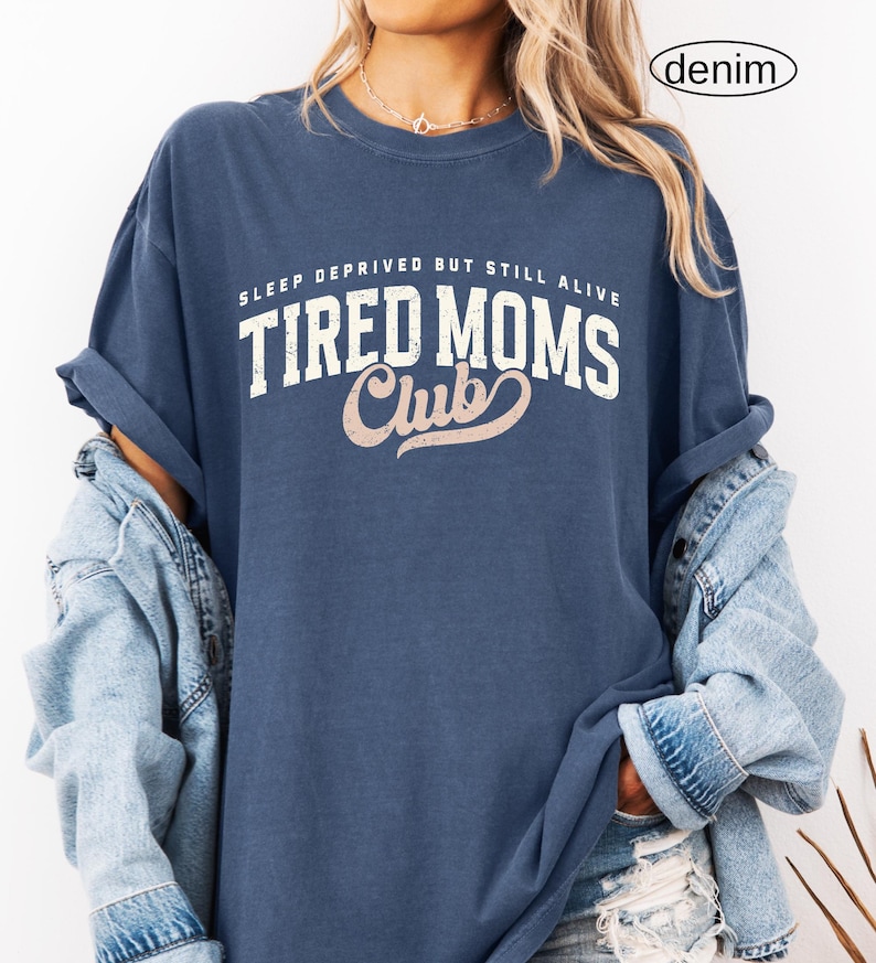 May include: A denim blue t-shirt with the text "SLEEP DEPRIVED BUT STILL ALIVE" above "TIRED MOMS Club" in white and pink lettering. The shirt has rolled-up sleeves and is paired with a denim jacket.