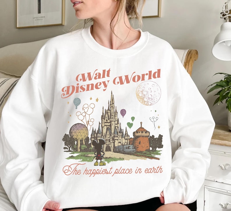 May include: White sweatshirt featuring a vintage-style graphic of Cinderella's castle, Mickey Mouse, and the text "Walt Disney World" and "The happiest place in earth". The design includes colorful balloons and a stylized moon.