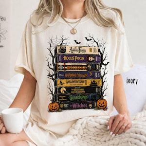 Op de afbeelding: Een ivoorkleurig t-shirt met een grafische print van een stapel VHS-banden met de titels The Addams Family, Hocus Pocus, Beetlejuice, Halloweentown, Halloweentown II, The Nightmare Before Christmas, Goosebumps en The Witches, omlijst door spinnenwebben en pompoenen.