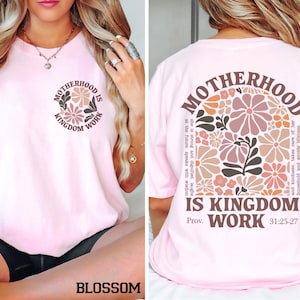 May include: A pale pink t-shirt featuring the text "MOTHERHOOD IS KINGDOM WORK" and a floral design. The back of the shirt has the same text and design. The shirt is a blossom colour.