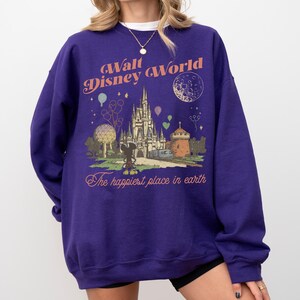 May include: A purple sweatshirt featuring a graphic of Cinderella's castle, Mickey Mouse, and the text "Walt Disney World" and "The happiest place on earth." The design includes balloons and a crescent moon.