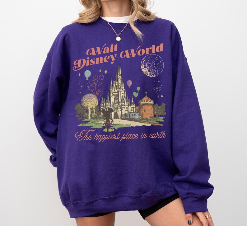 May include: A purple sweatshirt featuring a graphic of Cinderella's castle, Mickey Mouse, and the text "Walt Disney World" and "The happiest place on earth." The design includes balloons and a crescent moon.