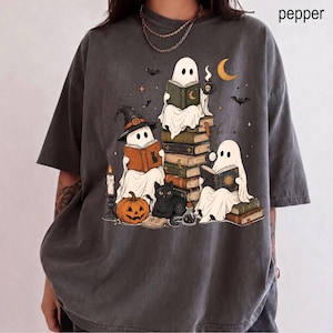 May include: A charcoal gray t-shirt featuring a graphic print of three white ghosts reading books. One ghost wears a witch hat, another sits on a stack of books, and the third holds a magnifying glass. A black cat, a pumpkin, and a candle are included.