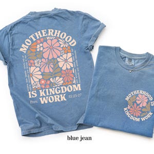 May include: Two blue jean-coloured t-shirts. One shirt features a large floral design with the words "MOTHERHOOD IS KINGDOM WORK" and a Bible verse reference. The other shirt has a smaller version of the design. A delicate necklace is on the second shirt.
