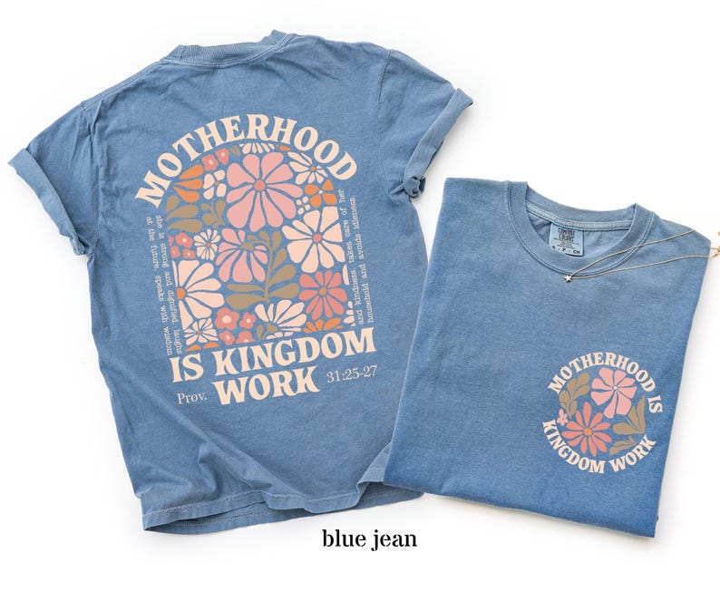 May include: Two blue jean-coloured t-shirts. One shirt features a large floral design with the words "MOTHERHOOD IS KINGDOM WORK" and a Bible verse reference. The other shirt has a smaller version of the design. A delicate necklace is on the second shirt.