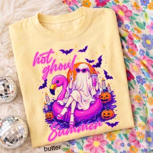 May include: Butter-colored t-shirt featuring a cartoon ghost wearing sunglasses, sitting on a pink flamingo float, and holding a drink. The shirt has the words "hot ghoul summer" in pink and purple, with bats and pumpkins.