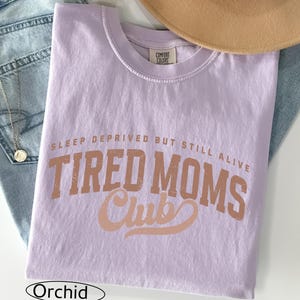 May include: A lavender-colored t-shirt with the text "SLEEP DEPRIVED BUT STILL ALIVE TIRED MOMS Club" in a distressed, brown font. The shirt is folded and rests on a pair of blue jeans. A tan hat is visible in the background.