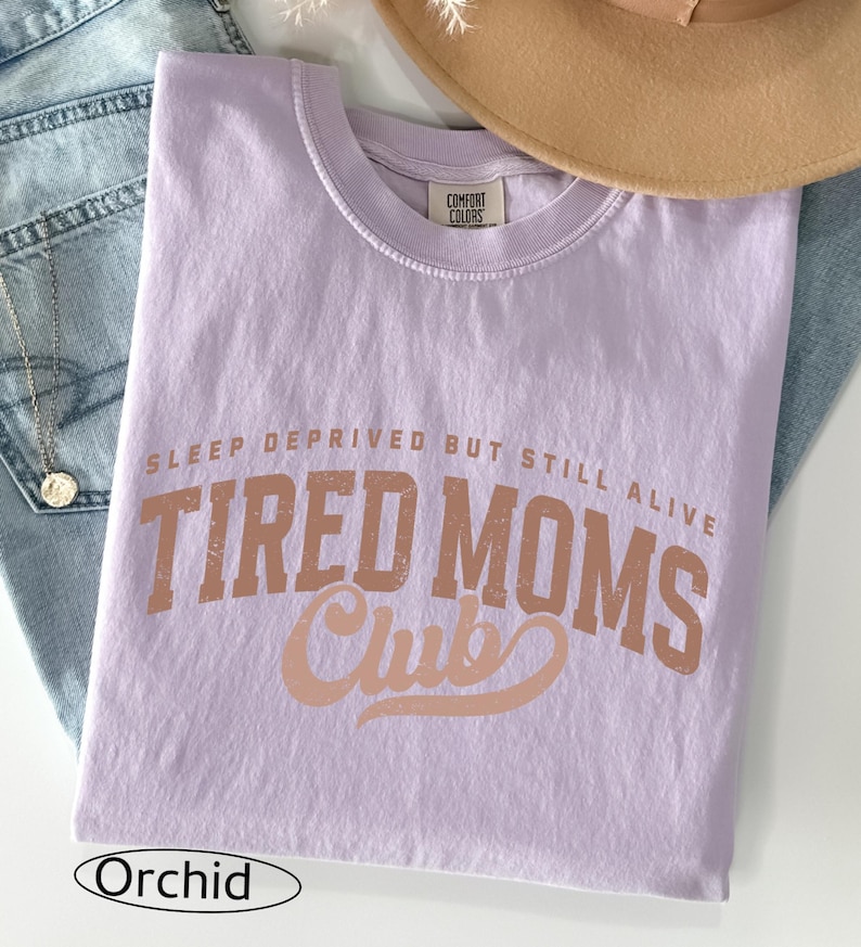 May include: A lavender-colored t-shirt with the text "SLEEP DEPRIVED BUT STILL ALIVE TIRED MOMS Club" in a distressed, brown font. The shirt is folded and rests on a pair of blue jeans. A tan hat is visible in the background.