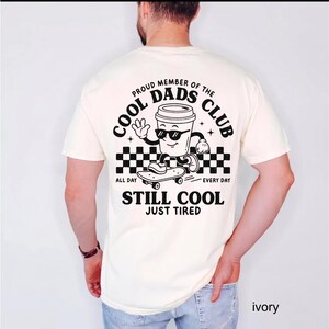Pu&ograve; includere: T-shirt color avorio con grafica in bianco e nero di una tazza di caff&egrave; con occhiali da sole su uno skateboard. Il testo recita "PROUD MEMBER OF THE COOL DADS CLUB" e "STILL COOL JUST TIRED".