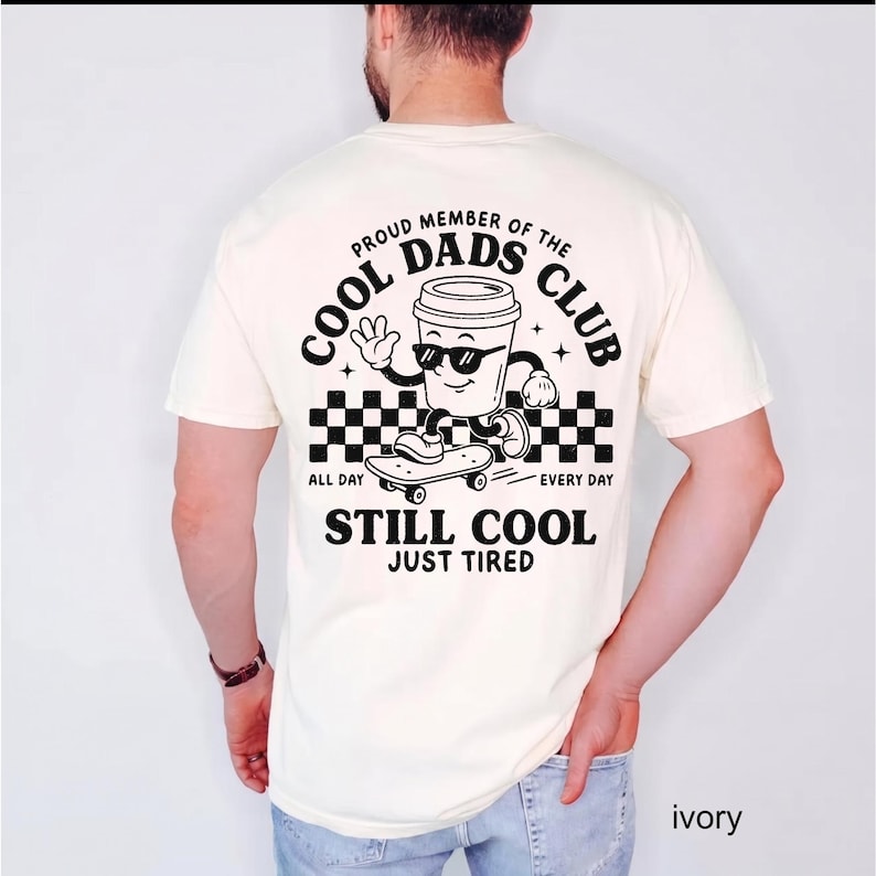 Pu&ograve; includere: T-shirt color avorio con grafica in bianco e nero di una tazza di caff&egrave; con occhiali da sole su uno skateboard. Il testo recita "PROUD MEMBER OF THE COOL DADS CLUB" e "STILL COOL JUST TIRED".