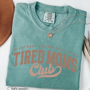 May include: Light green t-shirt with the text "SLEEP DEPRIVED BUT STILL ALIVE TIRED MOMS Club" in a brown font. A gold necklace with a pendant is on the shirt. The shirt is folded on a white surface.