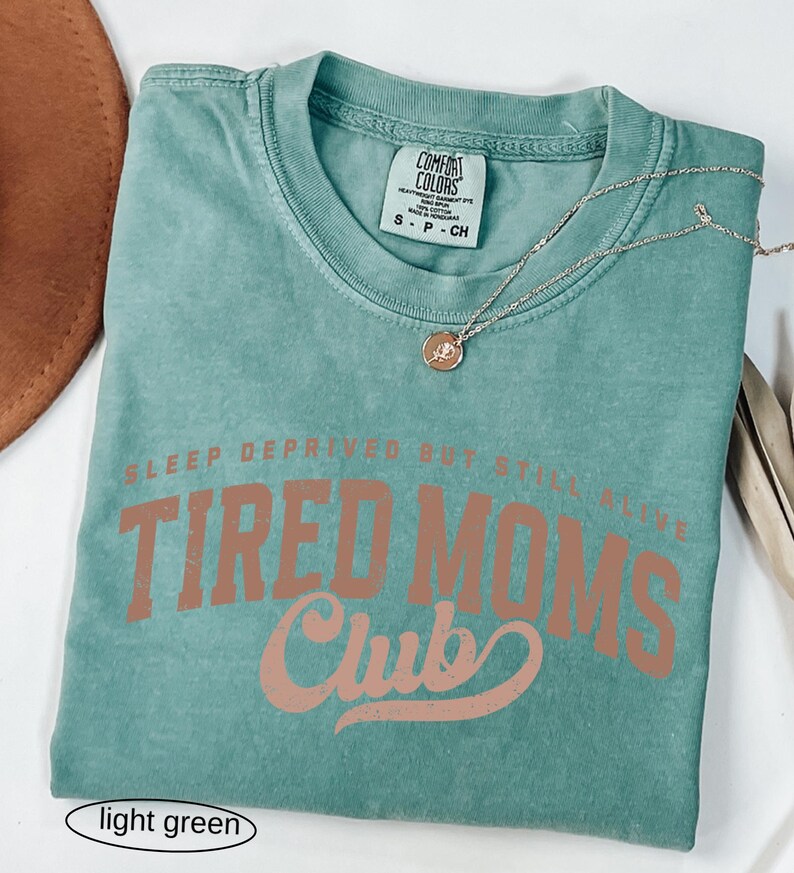 May include: Light green t-shirt with the text "SLEEP DEPRIVED BUT STILL ALIVE TIRED MOMS Club" in a brown font. A gold necklace with a pendant is on the shirt. The shirt is folded on a white surface.