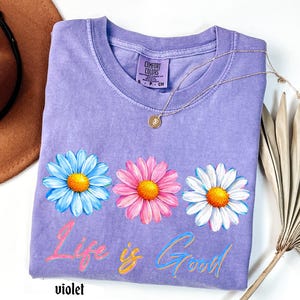 Pode incluir: Uma camiseta violeta Comfort Colors com tr&ecirc;s flores de margarida em azul, rosa e branco com centros amarelos. A frase "Life is Good" est&aacute; escrita em um script colorido abaixo das flores. Um colar de ouro repousa sobre a camisa.