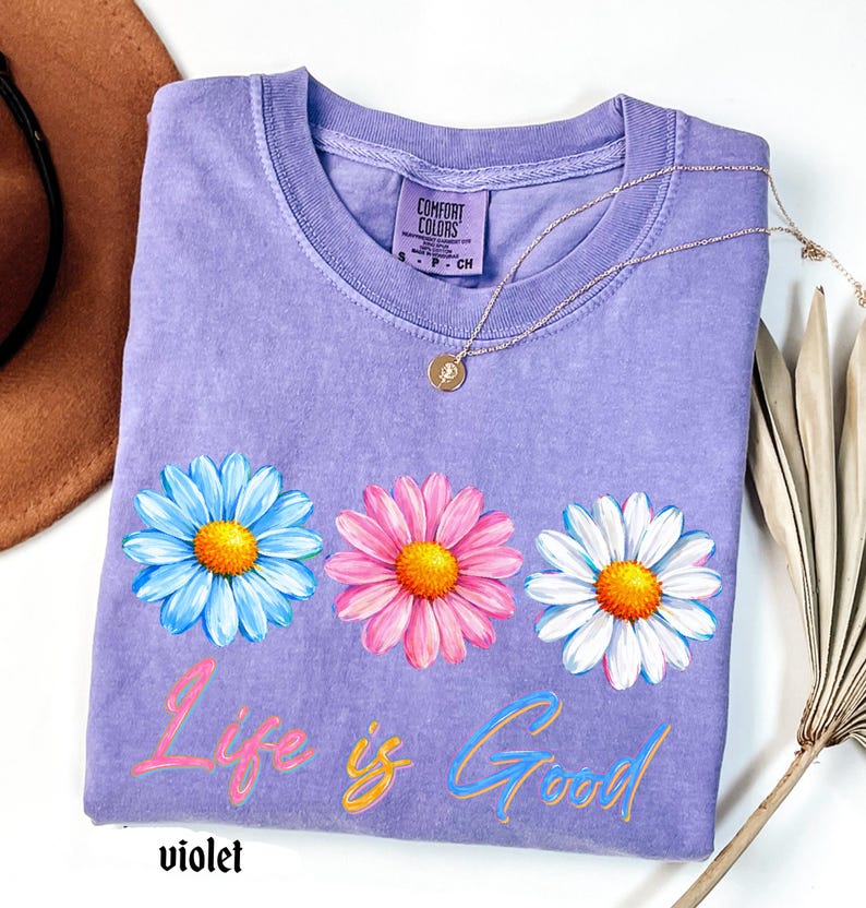 Pode incluir: Uma camiseta violeta Comfort Colors com tr&ecirc;s flores de margarida em azul, rosa e branco com centros amarelos. A frase "Life is Good" est&aacute; escrita em um script colorido abaixo das flores. Um colar de ouro repousa sobre a camisa.