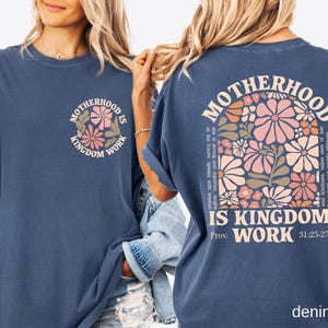 May include: A denim blue t-shirt featuring the text "MOTHERHOOD IS KINGDOM WORK" and a floral design on the front. The back of the shirt displays the same text and floral design, along with a quote from Proverbs 31:25-27.