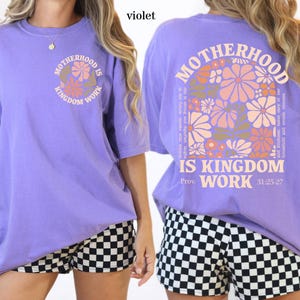 May include: A violet short-sleeved t-shirt with the text "MOTHERHOOD IS KINGDOM WORK" in a circular design on the front. The back of the shirt features the same text with floral graphics and the Bible verse Proverbs 31:25-27.