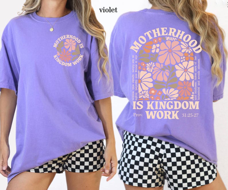 May include: A violet short-sleeved t-shirt with the text "MOTHERHOOD IS KINGDOM WORK" in a circular design on the front. The back of the shirt features the same text with floral graphics and the Bible verse Proverbs 31:25-27.