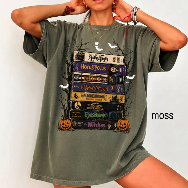 Op de afbeelding: Een mosgroen t-shirt met een grafische print van een stapel VHS-banden met titels zoals The Addams Family, Hocus Pocus, Beetlejuice, Halloweentown, The Nightmare Before Christmas, Goosebumps en The Witches, geflankeerd door twee pompoenen.