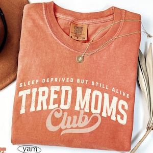 May include: A coral-colored t-shirt with the text "SLEEP DEPRIVED BUT STILL ALIVE TIRED MOMS Club" in white lettering. A gold necklace with a pendant rests on the shirt. The shirt has a Comfort Colors tag.