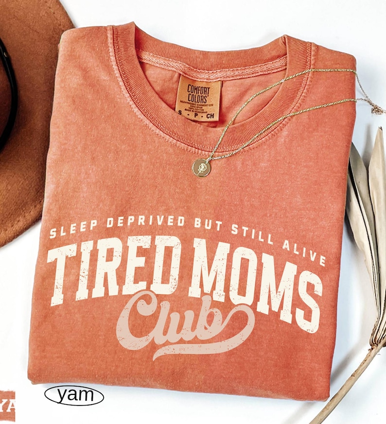 May include: A coral-colored t-shirt with the text "SLEEP DEPRIVED BUT STILL ALIVE TIRED MOMS Club" in white lettering. A gold necklace with a pendant rests on the shirt. The shirt has a Comfort Colors tag.