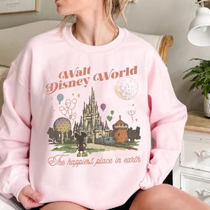 May include: Pink sweatshirt with a graphic of Cinderella's castle, Mickey Mouse, and the text "Walt Disney World" and "The happiest place in earth". The design includes balloons, a train, and a moon.