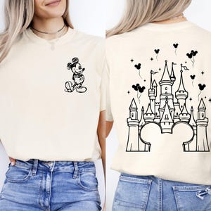 Puede incluir: Camiseta color crema con un boceto negro de Mickey Mouse en la parte delantera y un dise&ntilde;o de castillo en la parte trasera. El dise&ntilde;o del castillo incluye orejas de Mickey Mouse en la base, con globos y estrellas.