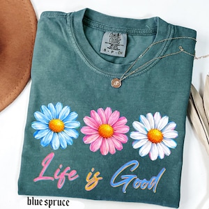 Pode incluir: Uma t-shirt azul-esverdeada com tr&ecirc;s desenhos de margaridas em azul, rosa e branco, cada uma com um centro amarelo. A frase "Life is Good" est&aacute; impressa abaixo das flores em letras coloridas. Um colar dourado est&aacute; sobre a t-shirt.