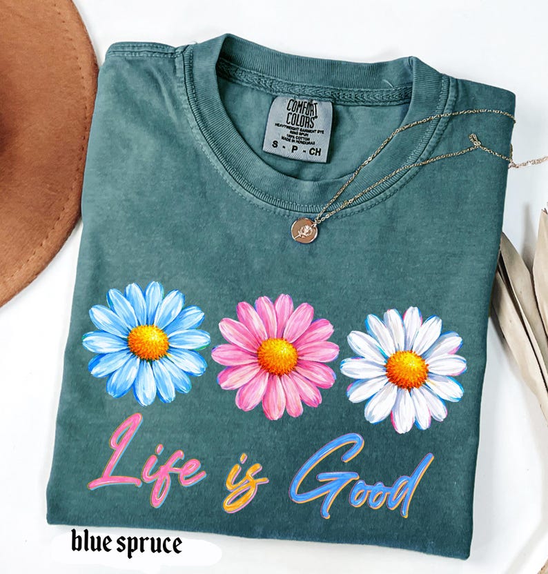 Pode incluir: Uma t-shirt azul-esverdeada com tr&ecirc;s desenhos de margaridas em azul, rosa e branco, cada uma com um centro amarelo. A frase "Life is Good" est&aacute; impressa abaixo das flores em letras coloridas. Um colar dourado est&aacute; sobre a t-shirt.