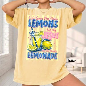 May include: Yellow t-shirt with a graphic of a glass of lemonade, lemons, and the text "Life Gave Me Some Lemons But My Jesus He Be Makin' Lemonade". The shirt has short sleeves and a relaxed fit.