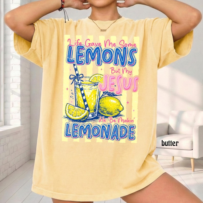 May include: Yellow t-shirt with a graphic of a glass of lemonade, lemons, and the text "Life Gave Me Some Lemons But My Jesus He Be Makin' Lemonade". The shirt has short sleeves and a relaxed fit.