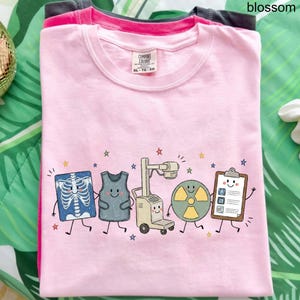 May include: A light pink crew neck t-shirt featuring a graphic print of five cartoon characters representing medical imaging equipment, including an X-ray, a lead apron, an X-ray machine, a radiation symbol, and a clipboard, surrounded by small stars.