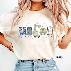 May include: An ivory t-shirt featuring a graphic print of five anthropomorphic medical items including an x-ray, a lead apron, an x-ray machine, a radiation symbol, and a clipboard, all accented with small colorful stars.