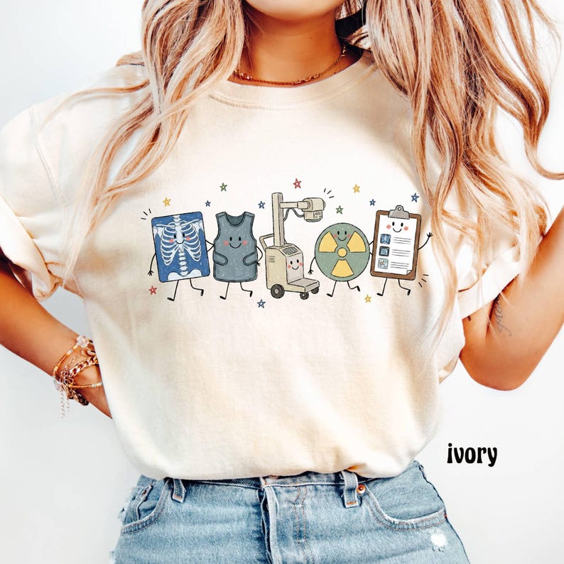 May include: An ivory t-shirt featuring a graphic print of five anthropomorphic medical items including an x-ray, a lead apron, an x-ray machine, a radiation symbol, and a clipboard, all accented with small colorful stars.