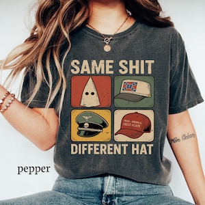 May include: Dark gray t-shirt with the text "SAME SHIT DIFFERENT HAT" and four images of hats. The images include a white hood, a Confederate flag hat, a military hat, and a red "Make America Great Again" hat.