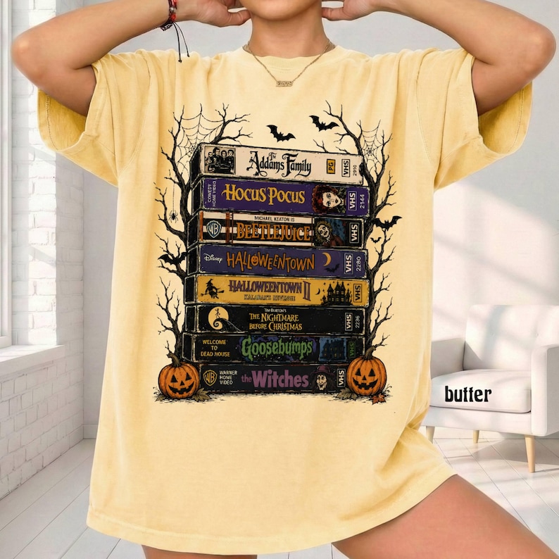 Op de afbeelding: Een lichtgeel T-shirt met korte mouwen en een grafische print van een stapel VHS-banden met de titels The Addams Family, Hocus Pocus, Beetlejuice, Halloweentown, Halloweentown II, The Nightmare Before Christmas, Goosebumps en The Witches.