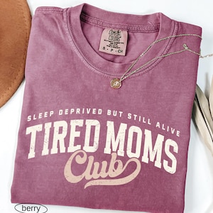 May include: A berry-colored Comfort Colors t-shirt with the text "SLEEP DEPRIVED BUT STILL ALIVE TIRED MOMS Club" in white and tan lettering. A gold-colored necklace with a small pendant rests on the shirt. A brown hat is visible in the upper left corner.