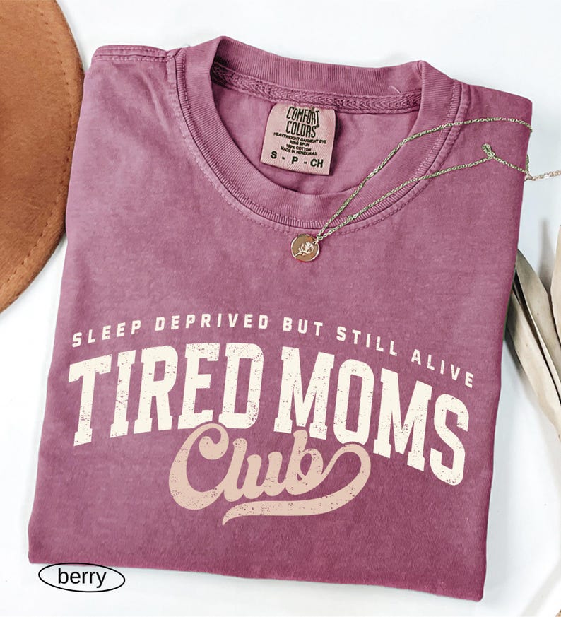 May include: A berry-colored Comfort Colors t-shirt with the text "SLEEP DEPRIVED BUT STILL ALIVE TIRED MOMS Club" in white and tan lettering. A gold-colored necklace with a small pendant rests on the shirt. A brown hat is visible in the upper left corner.