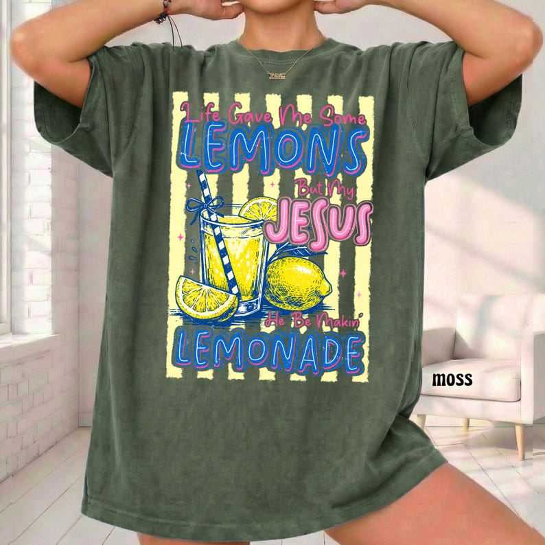 May include: Moss green t-shirt with a graphic of a lemonade drink, lemons, and the text "Life Gave Me Some Lemons But My Jesus He Be Makin' Lemonade". The design features yellow, blue, and pink colors.