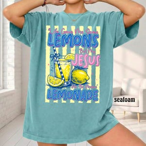May include: Seafoam green t-shirt with a graphic of a glass of lemonade, lemons, and the text "Life Gave Me Lemons But My Jesus Made Lemonade." The design has yellow and blue accents.