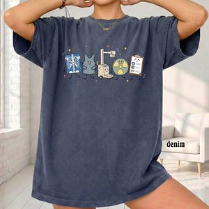 May include: A dark blue t-shirt featuring a graphic print of five anthropomorphic medical items: a chest x-ray, a scrub top, an x-ray machine, a radiation symbol, and a clipboard. Each item has a smiling face, thin black limbs, and small stars.