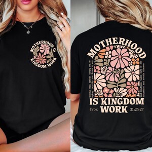 May include: Black t-shirt with the text "MOTHERHOOD IS KINGDOM WORK" in a floral design. The front of the shirt has a smaller version of the design. The back of the shirt features a larger floral design with the same text and Bible verse.