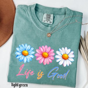 Pode incluir: Uma camiseta verde claro com tr&ecirc;s gr&aacute;ficos de flores de margarida em azul, rosa e branco, cada uma com um centro amarelo. O texto "Life is Good" &eacute; impresso abaixo das flores em rosa e azul. Um colar de prata com um pingente est&aacute; na camisa.