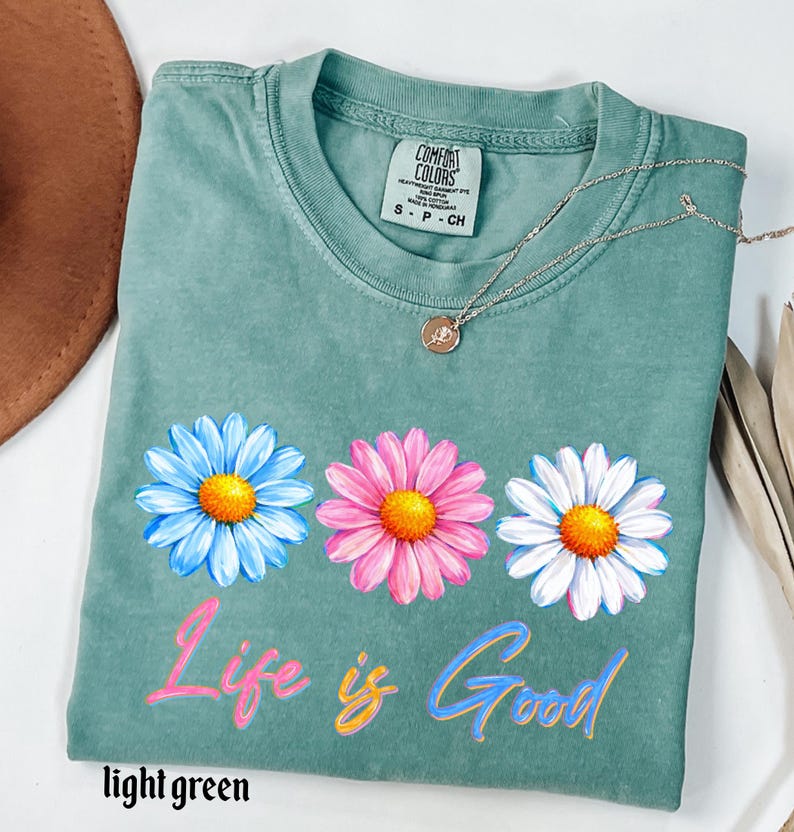 Pode incluir: Uma camiseta verde claro com tr&ecirc;s gr&aacute;ficos de flores de margarida em azul, rosa e branco, cada uma com um centro amarelo. O texto "Life is Good" &eacute; impresso abaixo das flores em rosa e azul. Um colar de prata com um pingente est&aacute; na camisa.