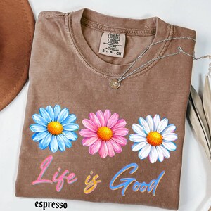 Pode incluir: Camiseta cor de caf&eacute; com tr&ecirc;s flores de margarida em azul, rosa e branco, cada uma com um centro amarelo. A frase "Life is Good" est&aacute; impressa abaixo das flores em rosa e azul. Um colar de prata com um pequeno pingente est&aacute; sobre a camiseta.