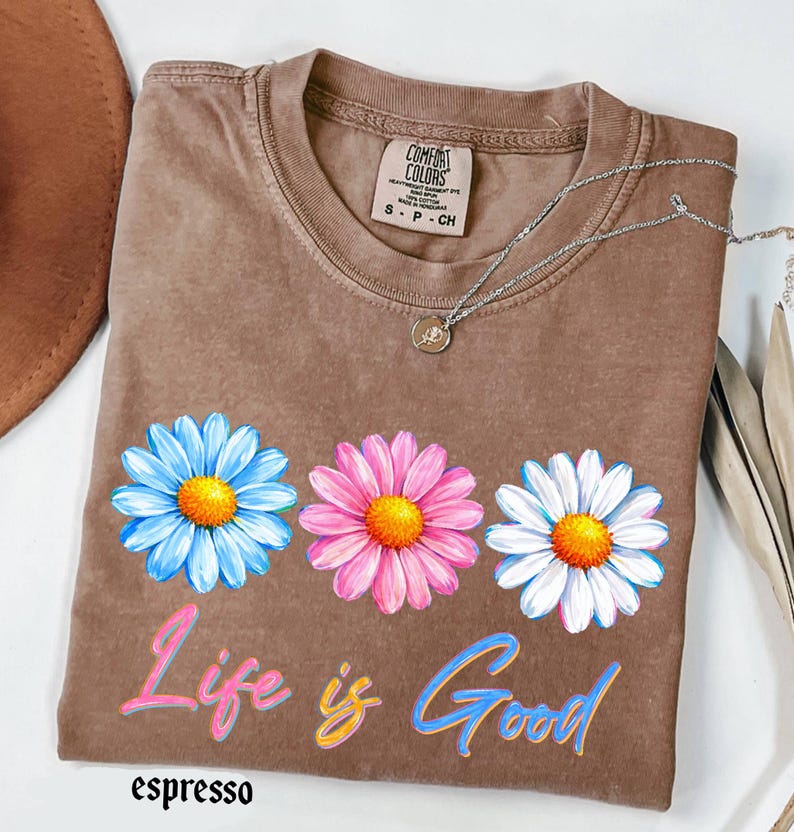 Pode incluir: Camiseta cor de caf&eacute; com tr&ecirc;s flores de margarida em azul, rosa e branco, cada uma com um centro amarelo. A frase "Life is Good" est&aacute; impressa abaixo das flores em rosa e azul. Um colar de prata com um pequeno pingente est&aacute; sobre a camiseta.