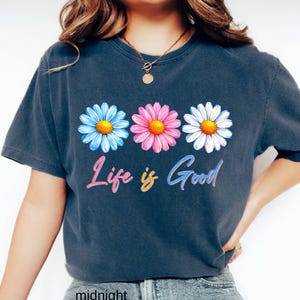 Pode incluir: Uma camiseta azul marinho com tr&ecirc;s flores de margarida em azul, rosa e branco com centros amarelos. A frase "Life is Good" est&aacute; escrita em uma fonte cursiva colorida abaixo das flores. A camiseta &eacute; de estilo casual.
