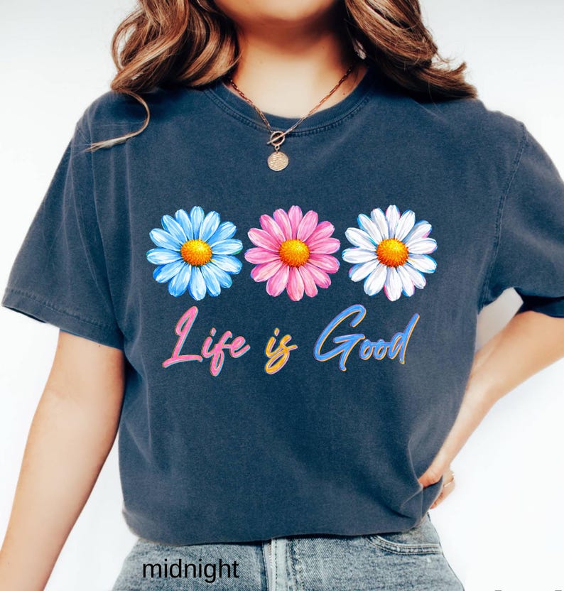 Pode incluir: Uma camiseta azul marinho com tr&ecirc;s flores de margarida em azul, rosa e branco com centros amarelos. A frase "Life is Good" est&aacute; escrita em uma fonte cursiva colorida abaixo das flores. A camiseta &eacute; de estilo casual.