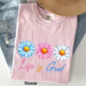 Pode incluir: Camiseta rosa claro com gola redonda e tr&ecirc;s margaridas azuis, rosas e brancas com centros amarelos. O texto "Life is Good" est&aacute; escrito em um script colorido. A palavra "blossom" est&aacute; escrita na parte inferior.